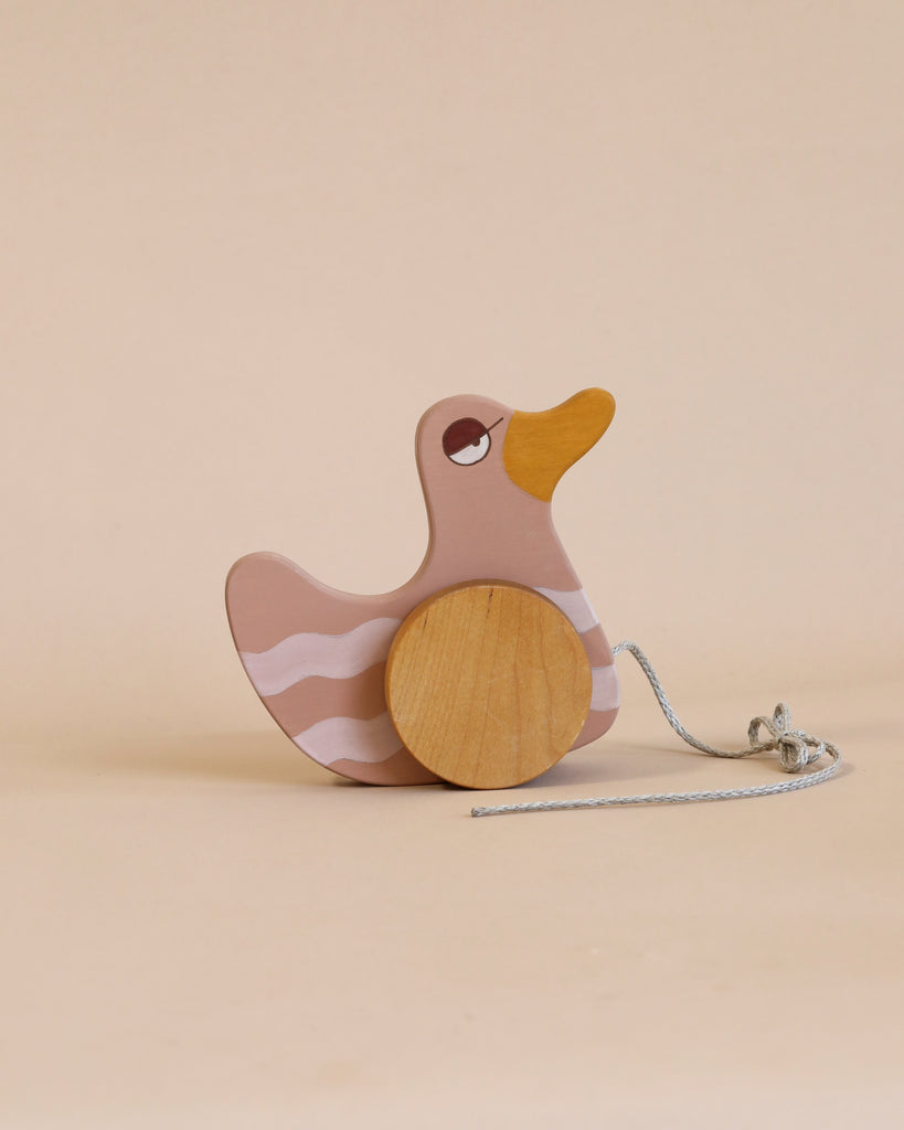A Duck Pull Toy - Pink with a pink and white body, an orange beak, and wooden wheels, standing on a beige background with a pull-string attached is crafted from sustainably harvested birch wood.