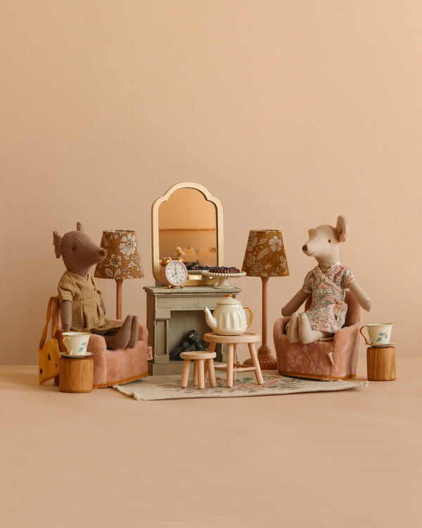 Two plush mice, including Maileg Mum Mouse, relax on armchairs in a pastel dollhouse living room with lamps and a fireplace, enjoying treats from the Maileg Moms Afternoon Tea Set—perfect for Mouse Paw Size fun.