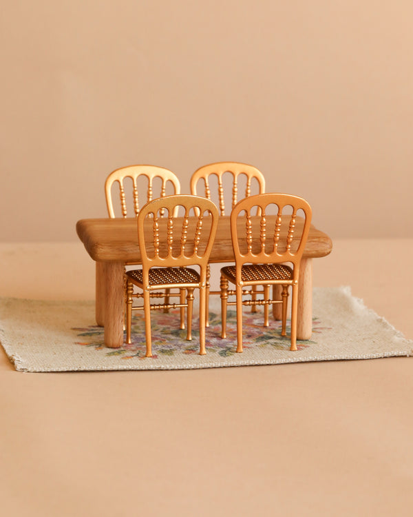 The Maileg Dining Room Set includes a petite wooden dollhouse table paired with four miniature gold chairs, all elegantly arranged on a flower rug against a soft beige background.