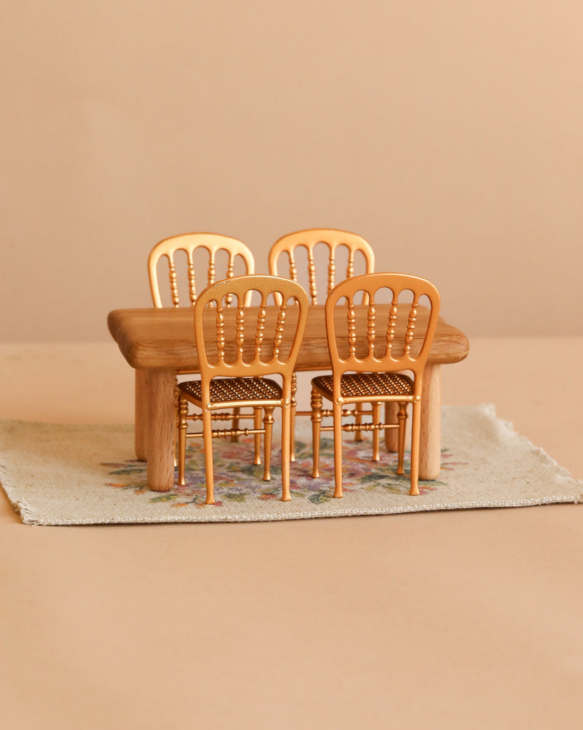 The Maileg Dining Room Set includes a petite wooden dollhouse table paired with four miniature gold chairs, all elegantly arranged on a flower rug against a soft beige background.