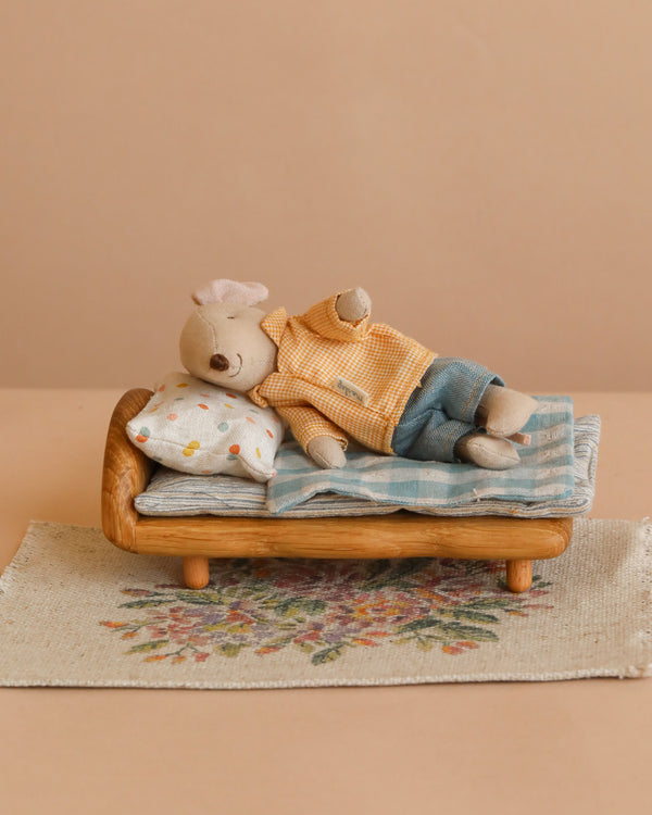 A small stuffed animal is lying on the Maileg Big Brother Bedtime Set, which includes a wooden dollhouse bed with a pillow and blanket. The bed sits on a flower rug, all set against a beige background.