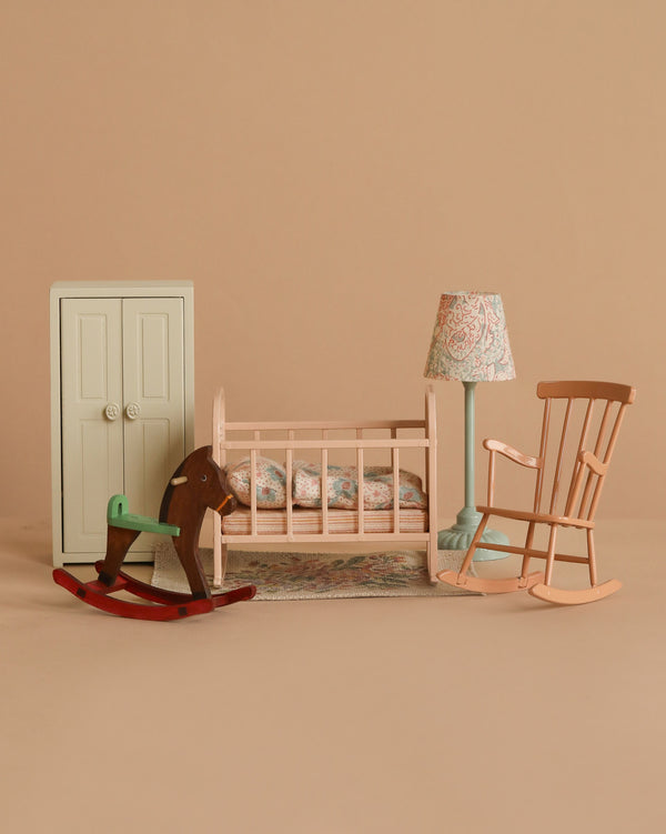 The Maileg Nursery Set includes miniature furniture for your Maileg Mouse: a white wardrobe, pink crib with bedding, green rocking horse, blue floor lamp with patterned shade, patterned rug, and pink rocking chair on a beige background.