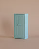 A small, light blue cabinet with double doors and round handles stands against a plain beige background, perfect for storing the Maileg Little Sister Bedtime Set or pairing with other Mouse Furniture.