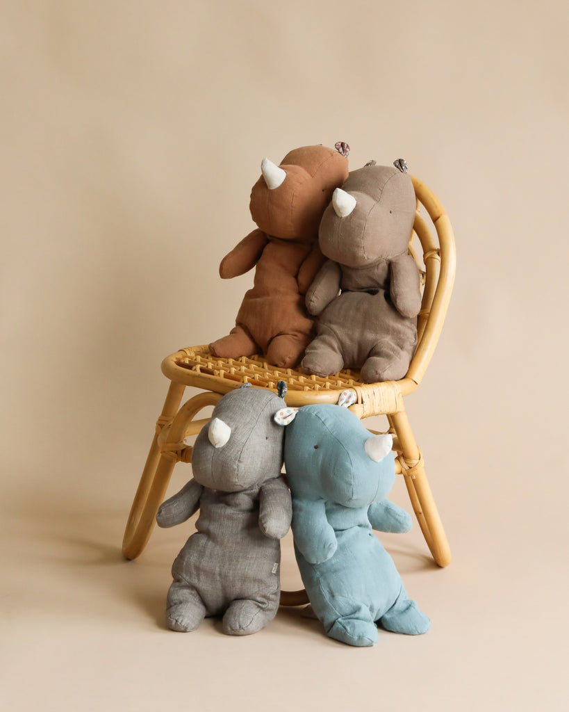 Four Maileg Medium Rhino plush toys provide cozy hugs around a wicker chair. A brown and a gray rhino sit on the chair like safari companions, while gray and blue rhinos lean against it. The background is neutral beige.