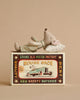 A Maileg Little Brother Mouse, wearing pajamas, rests on a pillow atop a vintage-style "Mouse Race" matchbox with race car art. Decorated with text and stars, this whimsical item enhances any display against a plain beige background.