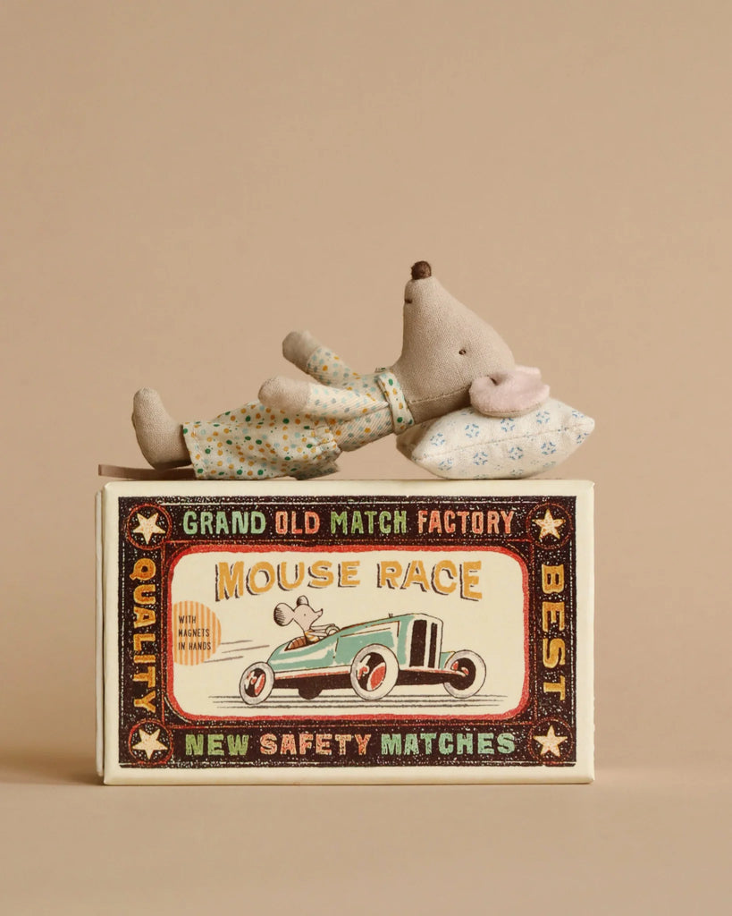 A Maileg Little Brother Mouse, wearing pajamas, rests on a pillow atop a vintage-style "Mouse Race" matchbox with race car art. Decorated with text and stars, this whimsical item enhances any display against a plain beige background.