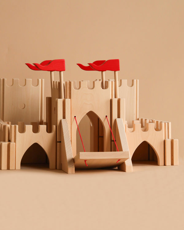 The Holztiger - Knight's castle is a handcrafted wooden European playset featuring red flags, a drawbridge, arched doorways, and battlements. Set against a beige background, its smooth construction makes it ideal for imaginative play.