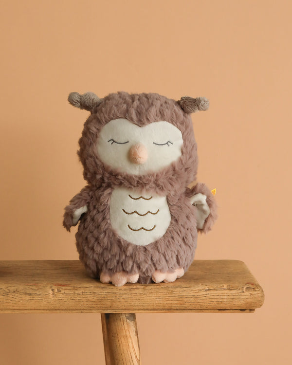 A Steiff Ollie Owl Stuffed Animal with closed eyes, a light beige face and belly, and a fluffy brown body sits on a wooden bench against a peach background, highlighting its premium plush softness.