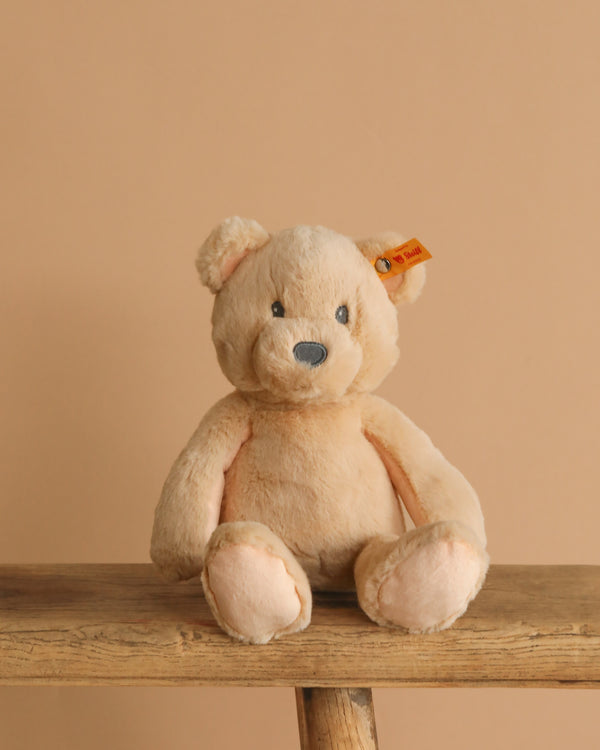 The Steiff Bearzy Teddy Bear, a light brown and cuddly companion with a small ear tag, sits on a wooden bench against a beige background.