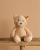 The Steiff Bearzy Teddy Bear, a light brown and cuddly companion with a small ear tag, sits on a wooden bench against a beige background.