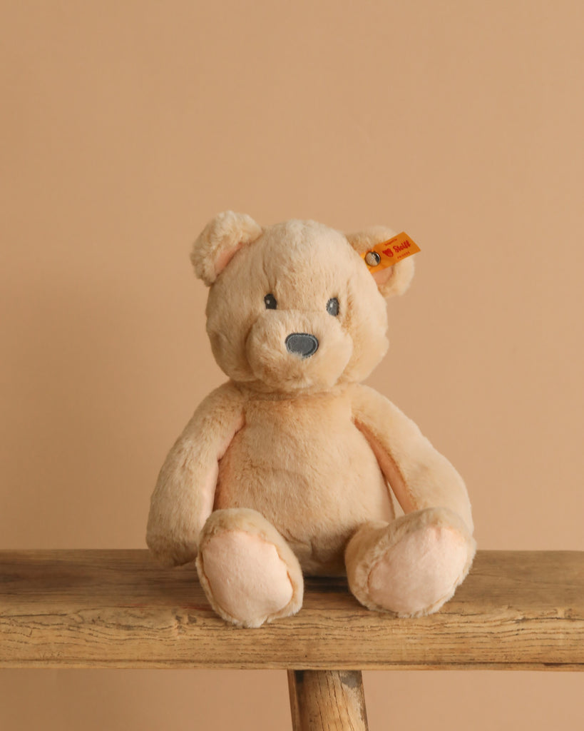 The Steiff Bearzy Teddy Bear, a light brown and cuddly companion with a small ear tag, sits on a wooden bench against a beige background.
