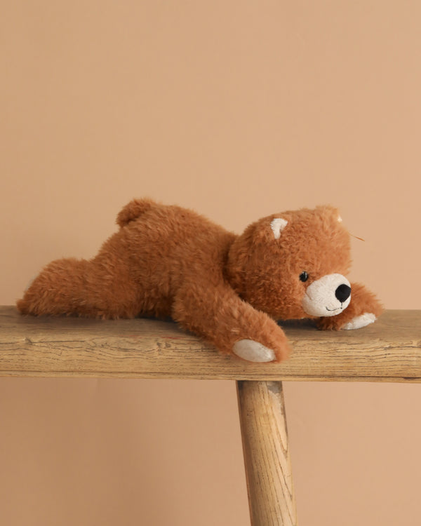 The Steiff Barny Brown Bear Stuffed Animal, a brown plush bear, lies on its stomach across a wooden bench against a plain beige background, evoking the charm of classic Steiff bears.