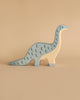 A wooden toy dinosaur with a long neck and tail, painted in a light blue color with darker blue crescent-shaped markings along its back, stands against a plain beige background. This handcrafted wood Holztiger Brontosaurus is stylized and simplistic in design, typical of HOLZTIGER figures made in Europe.