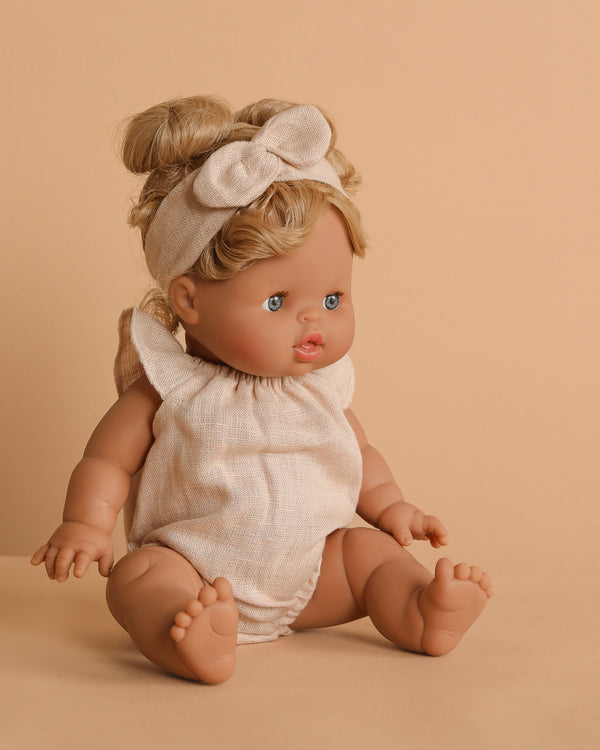 The Minikane Baby Doll (13") - Louise With Outfit, featuring lifelike details, blonde hair in a bun with a bow headband and a light beige romper, sits on a beige surface against a matching background.