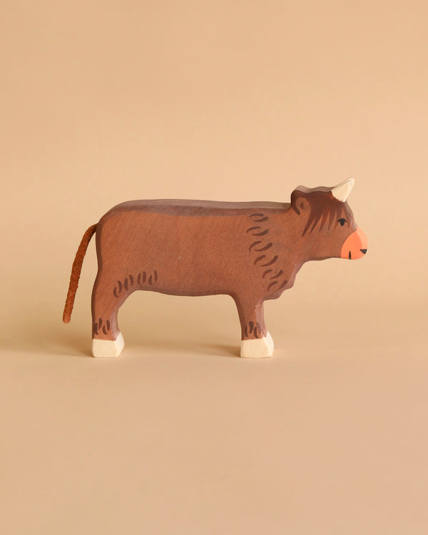 A wooden toy cow with a brown body, white hooves, a red nose, and tiny horns set against a beige background. Handcrafted out of maple and beech wood, this simplistic yet charming Holztiger Highland Cattle, Standing figure boasts minimal detailing for a rustic appearance. Made in Europe to ensure high quality craftsmanship.