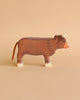 A wooden toy cow with a brown body, white hooves, a red nose, and tiny horns set against a beige background. Handcrafted out of maple and beech wood, this simplistic yet charming Holztiger Highland Cattle, Standing figure boasts minimal detailing for a rustic appearance. Made in Europe to ensure high quality craftsmanship.