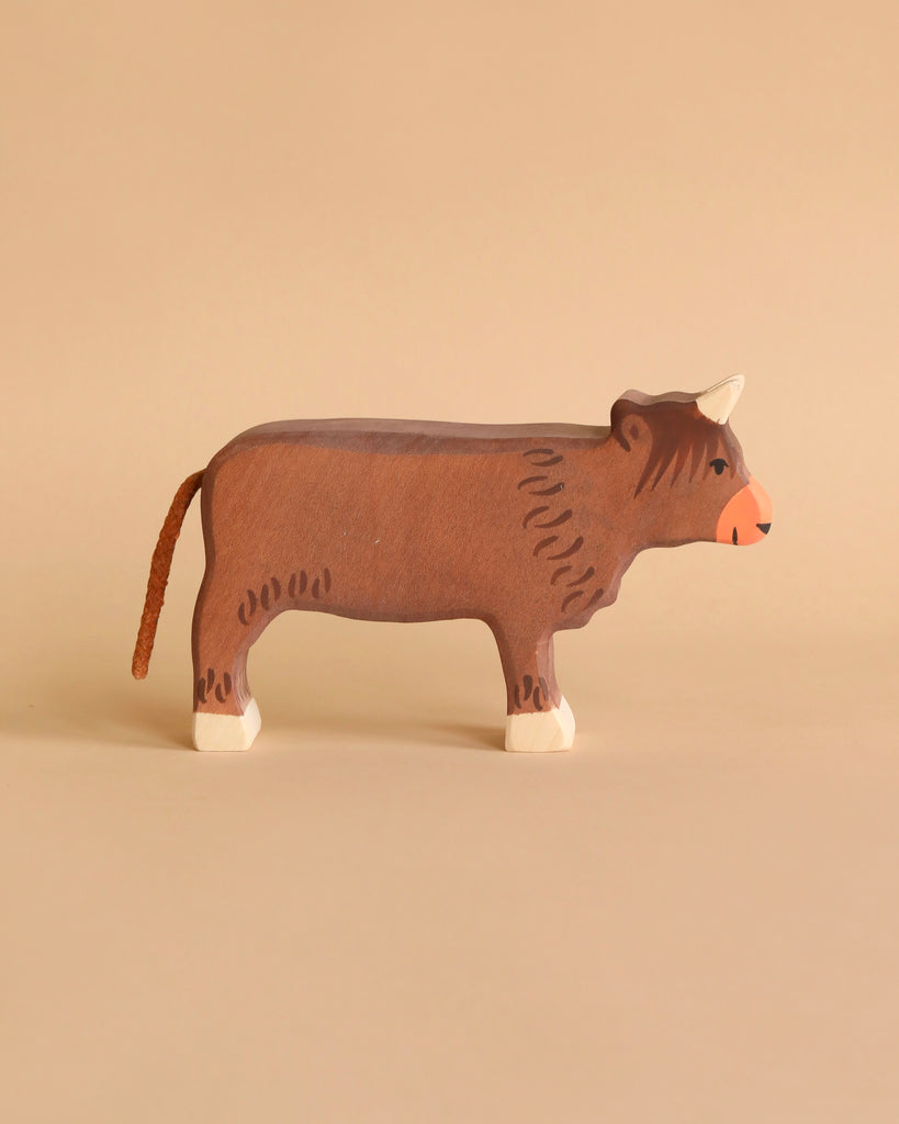 A wooden toy cow with a brown body, white hooves, a red nose, and tiny horns set against a beige background. Handcrafted out of maple and beech wood, this simplistic yet charming Holztiger Highland Cattle, Standing figure boasts minimal detailing for a rustic appearance. Made in Europe to ensure high quality craftsmanship.