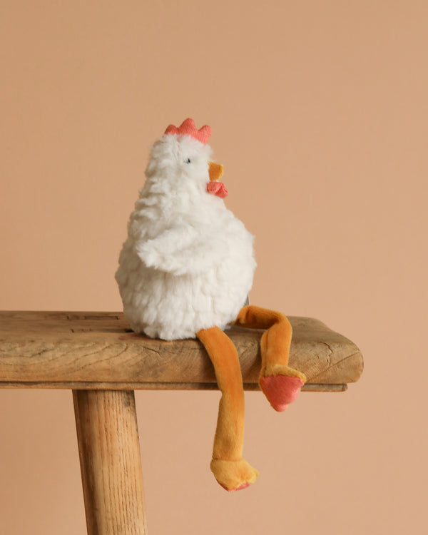 Charlie the Chicken Rattle Stuffed Animal, a soft white plush toy with orange legs and beak, sits on a rustic wooden bench against a beige background.