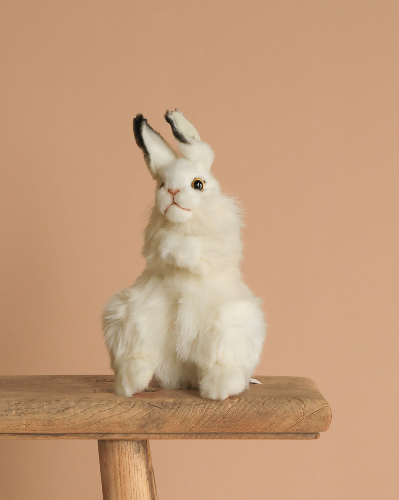 The White Bunny Stuffed Animal, featuring hand-sewn realistic details and black-tipped ears, sits upright on a rustic wooden stool against a plain beige background.