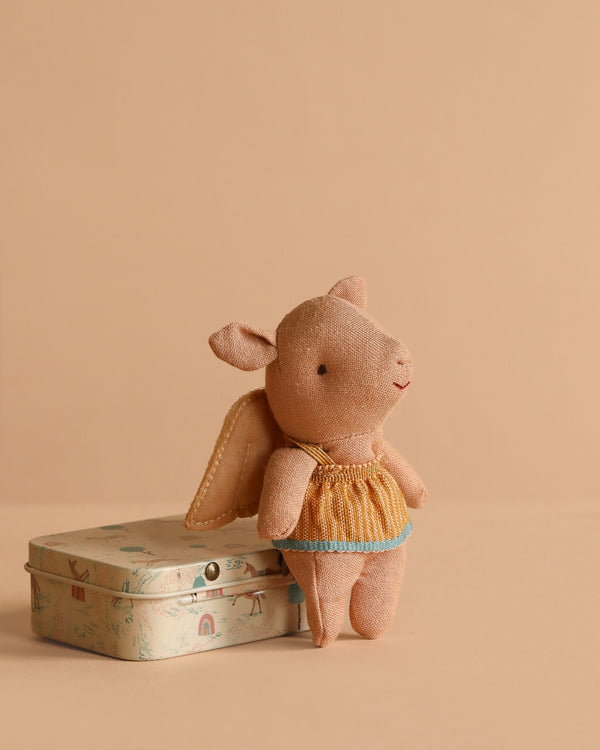 The Maileg Pig In A Box With Fairy Outfit, dressed in yellow and blue fairy clothes, stands beside a playful tin on a beige background—an adorable gift for any baby girl.