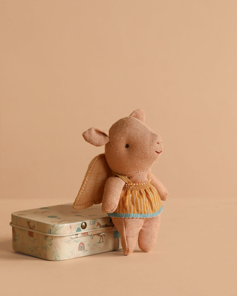 The Maileg Pig In A Box With Fairy Outfit, dressed in yellow and blue fairy clothes, stands beside a playful tin on a beige background—an adorable gift for any baby girl.