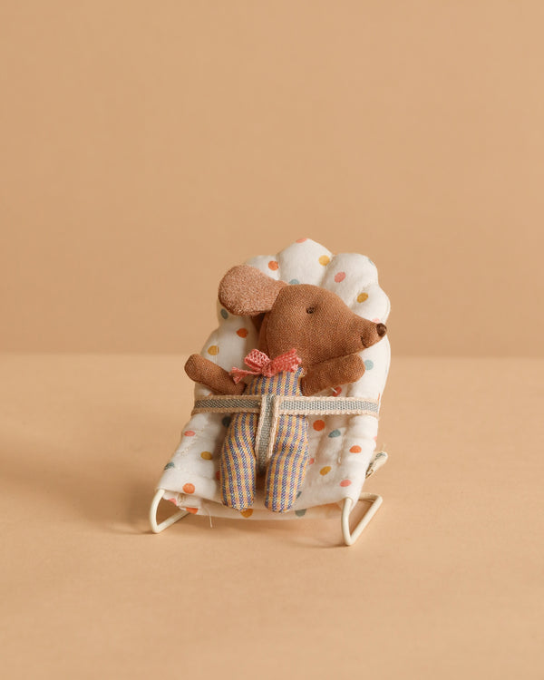 The Maileg Sleepy Wakey Baby Mouse In Bouncer wears striped pants and a red bow, sitting in a soft white polka-dotted bouncer against a beige background.