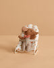 The Maileg Sleepy Wakey Baby Mouse In Bouncer wears striped pants and a red bow, sitting in a soft white polka-dotted bouncer against a beige background.