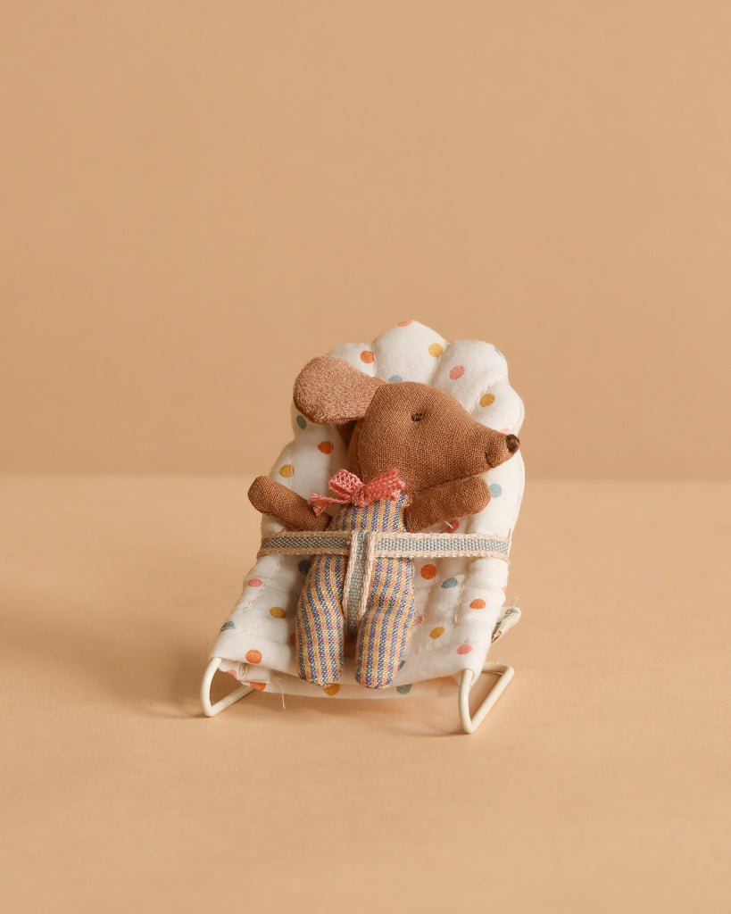 The Maileg Sleepy Wakey Baby Mouse In Bouncer wears striped pants and a red bow, sitting in a soft white polka-dotted bouncer against a beige background.