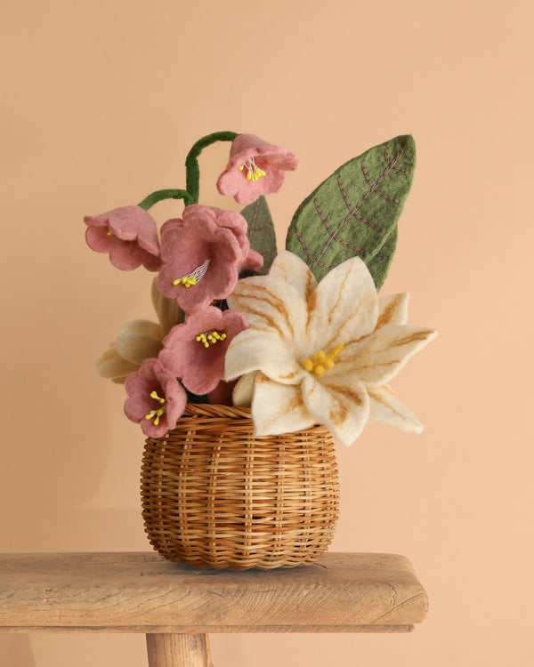 The Handcrafted Felt Flower Bouquet, featuring cream lilies, pink blooms, and green leaves in a small wicker basket, is displayed on a wooden surface against a beige background.