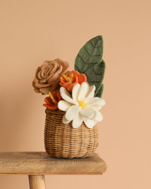 The Handcrafted Felt Flower Bouquet features brown, orange, and white felt flowers with green leaves in a small wicker basket. Displayed on a wooden stool against a beige background, these sustainable florals add charm to any space.