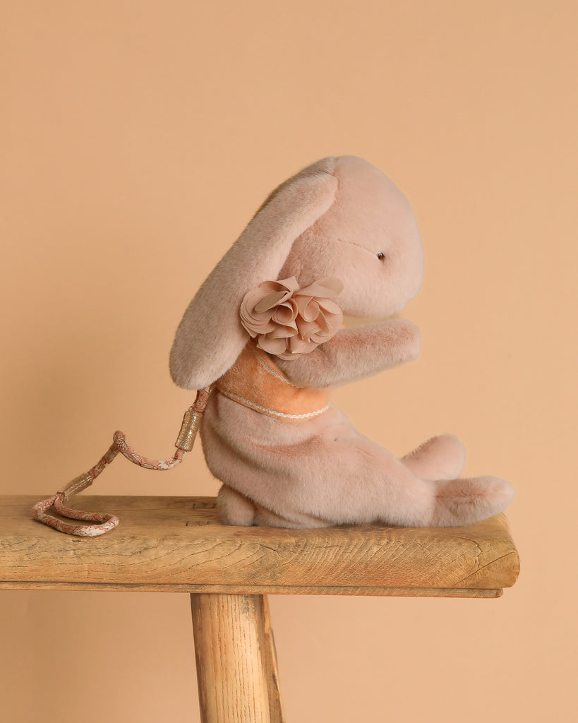 A soft, light pink Maileg Plush Sleeping Bunny With Harness sits on a wooden bench against a beige background.