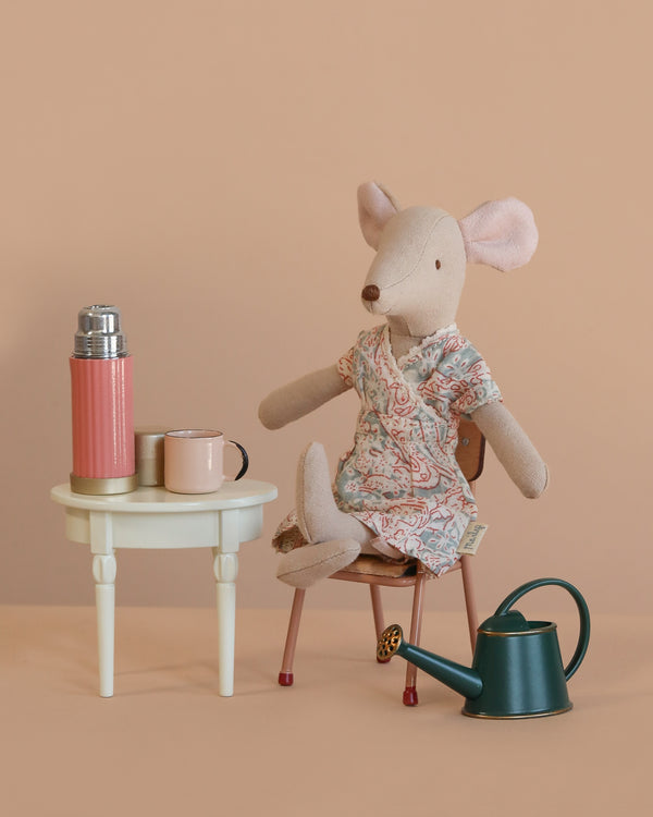 The Maileg Mom Mouse Garden Relaxation Set features a mom mouse in a floral dress on a chair beside a mini side table with a pink thermos, mug, and watering can, all against a soft beige background.