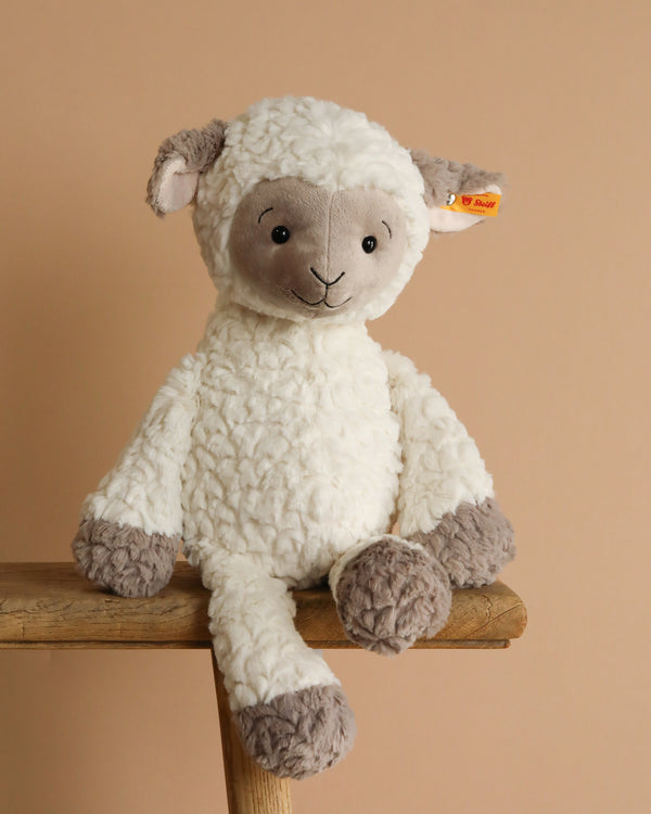 The Steiff Lita Lamb Stuffed Animal - 18" features a soft white, curly body with gray face, ears, and feet. Seated on a wooden stool against a beige background, this plush toy sheep with an orange tag is perfect as a children's gift.