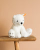 The Steiff Lasse Polar Bear Stuffed Animal - 11", crafted from ultra-soft white fabric and featuring a yellow ear tag, sits on a wooden bench against a plain beige background.