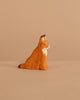 The Ostheimer Lynx Sitting, a small handcrafted wooden figurine painted orange with brown spots and a white chest, perfect for imaginative play, sits on a beige surface against a matching beige background.