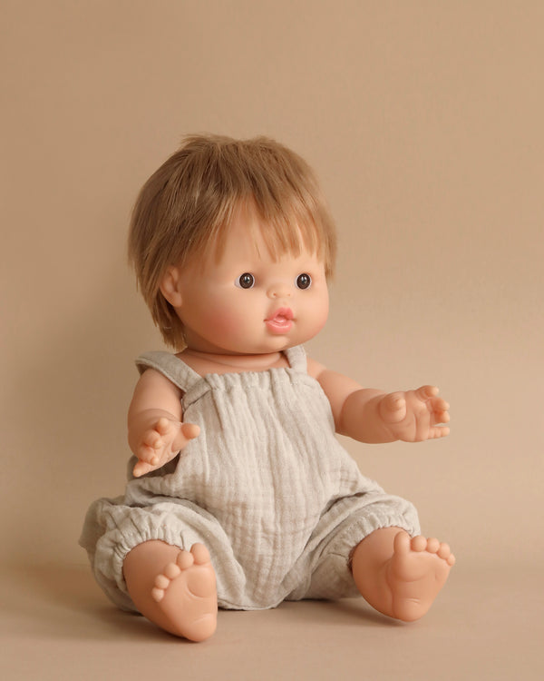 A small Minikane Baby Doll (13") - Achille with short, light brown hair is seated against a plain beige background. The anatomically correct doll is dressed in a light-colored, sleeveless romper with gathered cuffs at the legs. Its arms are slightly extended forward, and it has a neutral facial expression.