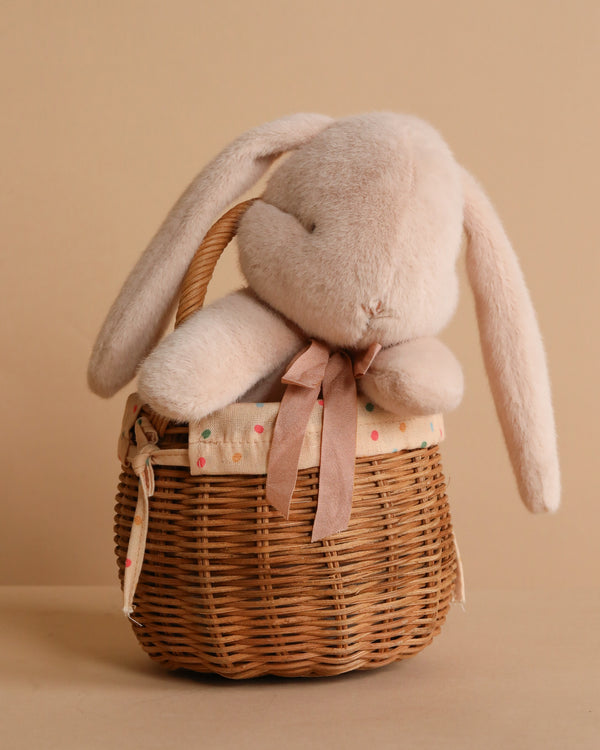 The Maileg Bunny And Basket Set features a soft beige bunny with floppy ears and a ribbon around its neck, sitting in a fabric-lined rattan basket against a plain tan background.