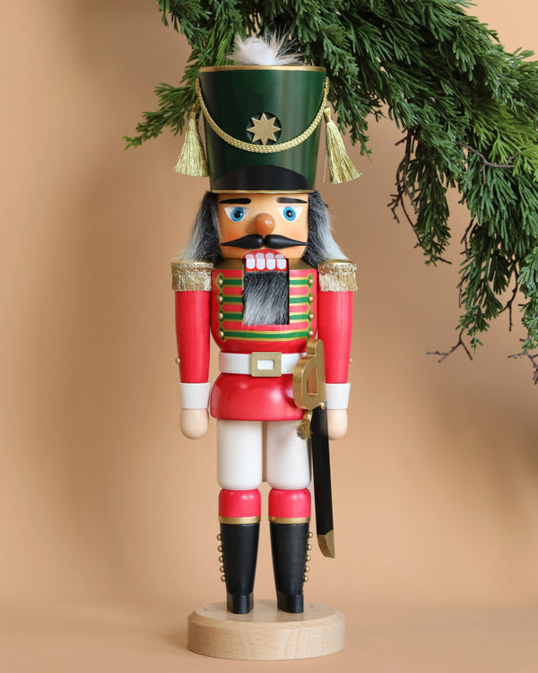 The Christian Ulbricht Collectible Wooden Nutcracker - Red Soldier stands upright against a beige background, reminiscent of traditional German-made nutcrackers. This wooden figure is outfitted in a red and white uniform with green accents and gold epaulettes, holding a sword as pine branches hang above it.