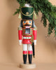 The Christian Ulbricht Collectible Wooden Nutcracker - Red Soldier stands upright against a beige background, reminiscent of traditional German-made nutcrackers. This wooden figure is outfitted in a red and white uniform with green accents and gold epaulettes, holding a sword as pine branches hang above it.