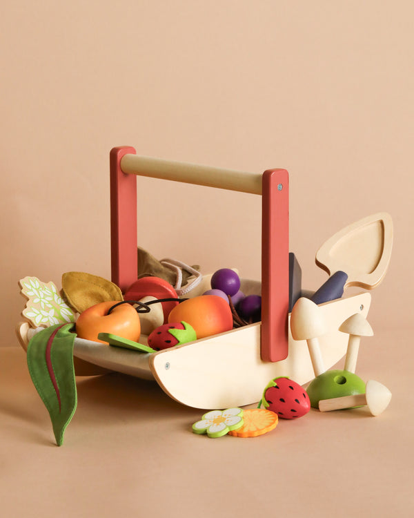 The Wild Wood Foraging Trug, a wooden toy basket with a red handle, is filled with colorful wooden fruits and vegetables for imaginative play; a mushroom and strawberry sit beside it on a beige background.