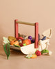 The Wild Wood Foraging Trug, a wooden toy basket with a red handle, is filled with colorful wooden fruits and vegetables for imaginative play; a mushroom and strawberry sit beside it on a beige background.