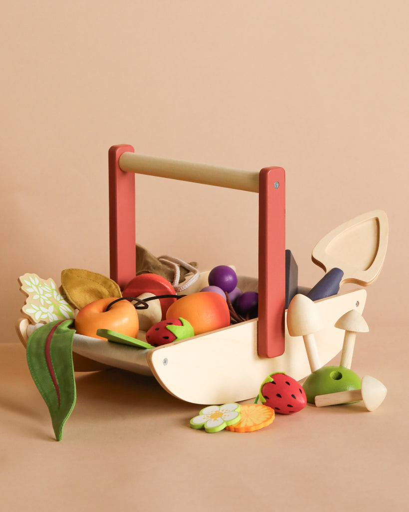 The Wild Wood Foraging Trug, a wooden toy basket with a red handle, is filled with colorful wooden fruits and vegetables for imaginative play; a mushroom and strawberry sit beside it on a beige background.