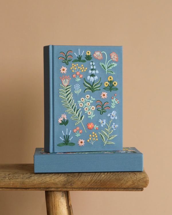 The Menagerie Garden Embroidered Journal, a blue hardcover with colorful floral embroidery, stands upright on a matching box atop a wooden stool against a plain beige background.