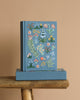 The Menagerie Garden Embroidered Journal, a blue hardcover with colorful floral embroidery, stands upright on a matching box atop a wooden stool against a plain beige background.