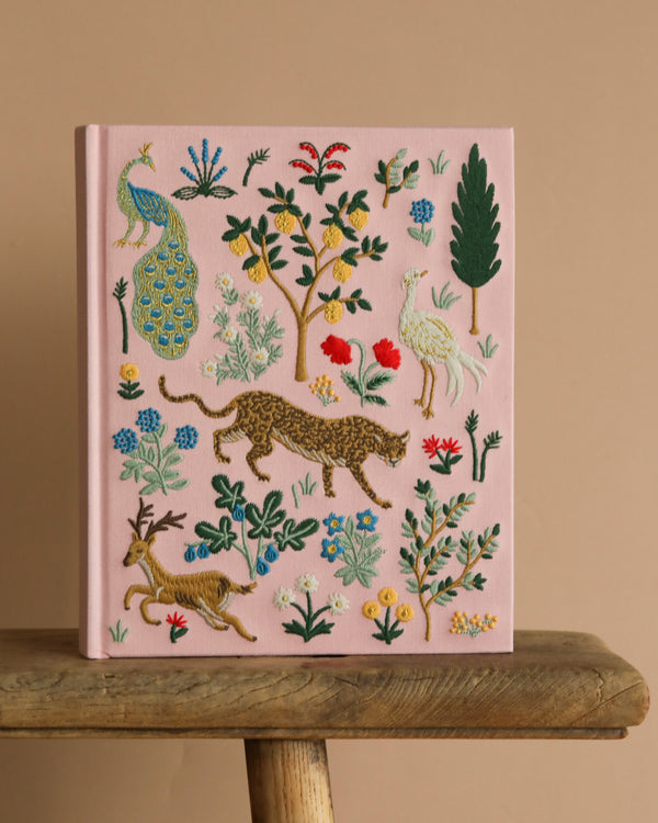 The Menagerie Embroidered Sketchbook, in pink with colorful animal and floral embroidery, sits on a wooden bench against a beige background.