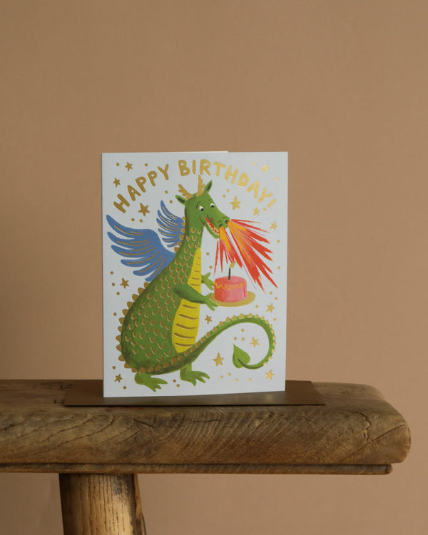The Birthday Dragon Card features a green winged dragon breathing fire onto a pink cake, surrounded by gold foil stars and "HAPPY BIRTHDAY!" text. Presented on a wooden surface with a beige background. Blank inside.