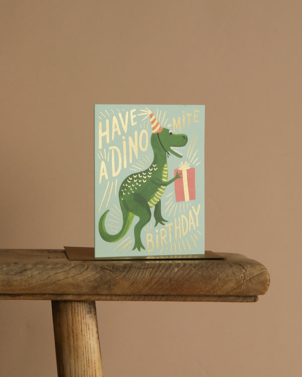The Dino-mite Birthday Card shows a green dinosaur in a party hat holding a gift, with "Have a dino-mite birthday" on a light green background. It comes with a metallic gold envelope and has a blank interior for your personal message.