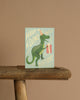 The Dino-mite Birthday Card shows a green dinosaur in a party hat holding a gift, with "Have a dino-mite birthday" on a light green background. It comes with a metallic gold envelope and has a blank interior for your personal message.