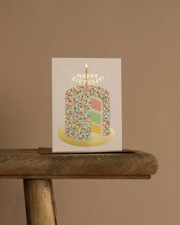 The Layer Cake Card features a sprinkle-topped rainbow cake with a lit candle, set on a wooden stool against a beige background. It includes metallic gold foil accents and has a blank interior for your personal message.