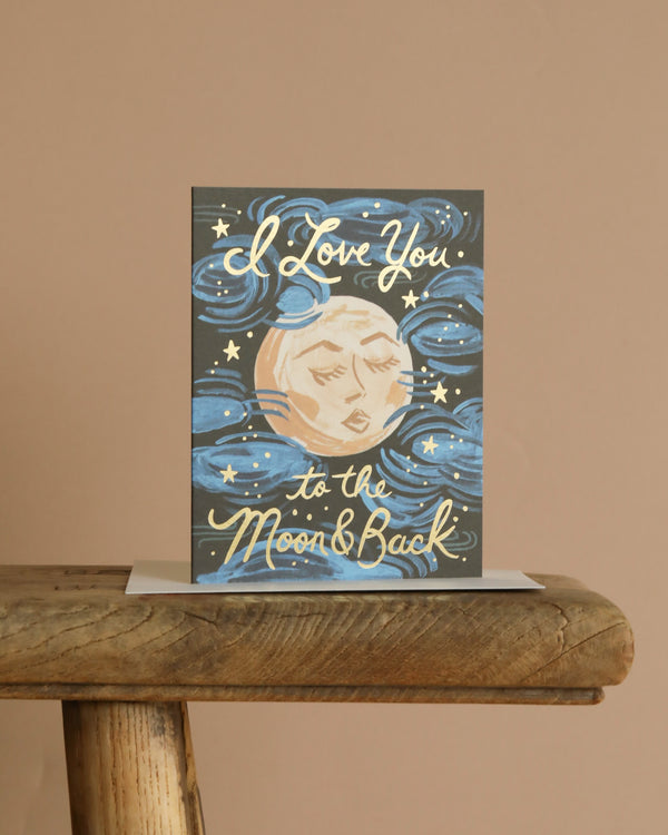 The To the Moon and Back Card features an illustrated moon with a face, blue swirls, stars, and gold foil accents highlighting "I Love You to the Moon & Back." Blank inside for your message. Displayed on a wooden surface.
